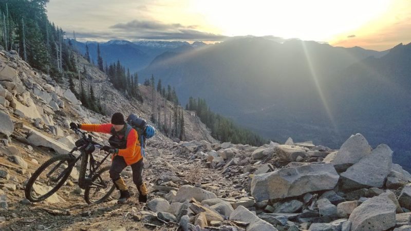 bikerumor pic of the day man in orange jacket pushing mountain bike up rocky mountain as sun rises behind him.