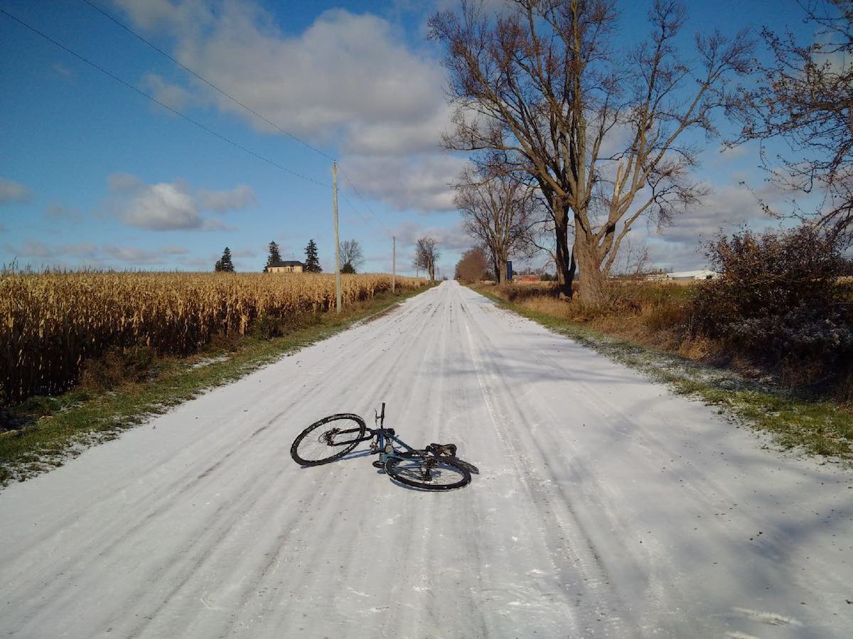 bikerumor pic of the day black bicycle laying on gravel road with dried corn fields on one side and blue skies ahead north of Woodstock, Ontario.