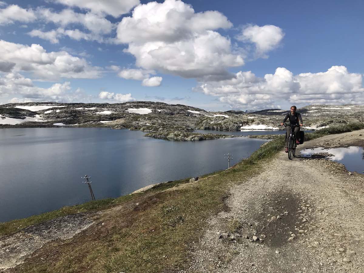 bikerumor pic of the day cyclist riding on the edge of a canal on the Rallarvegen of Norway blue skies dotted with clouds.