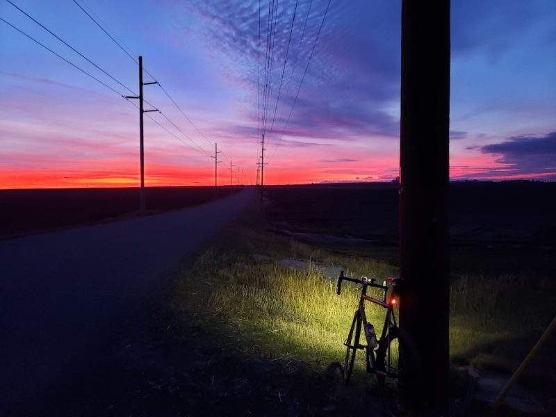 Bikerumor pic of the day of a bicycle leaning against a telephone pole with light beam in the grass and orange and blue sunset in the distance.