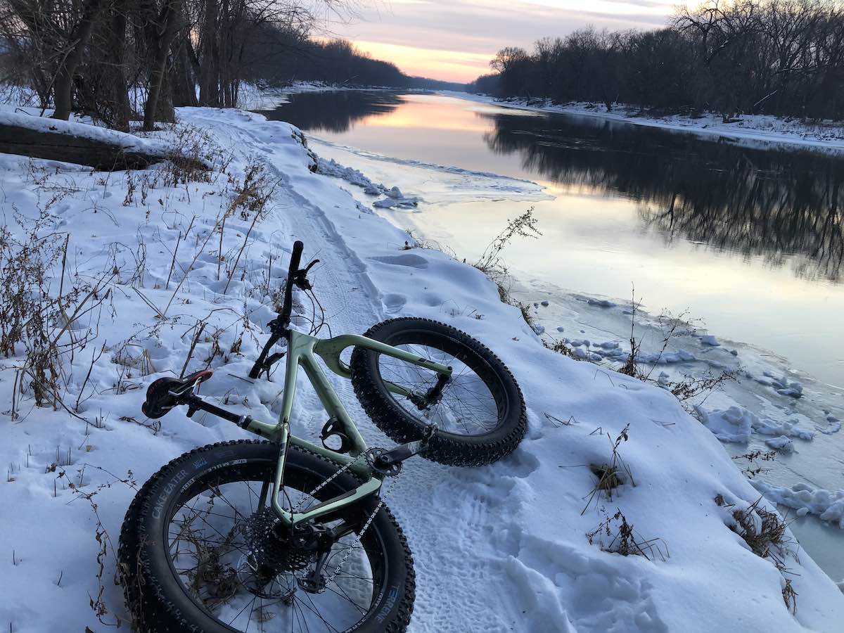 bikerumor pic of the day green fat bike laying in the snow alongside the river on the Minnesota River Trail in Bloomington, Minnesota. At sunset with light orange glow in the sky in the distance.