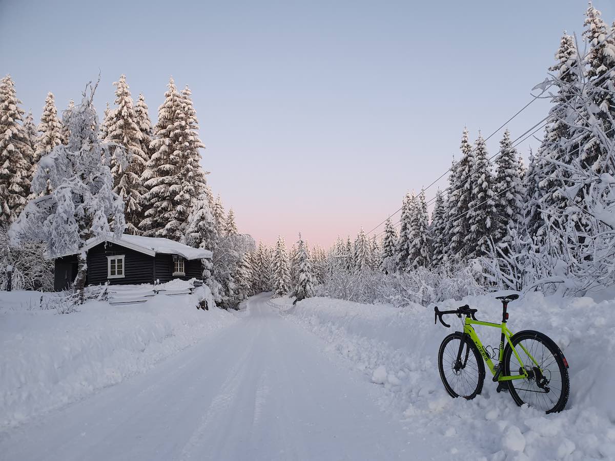 bikerumor pic of the day bicycle on the right of a snow covered road, with snow on surrounding trees and an orange glow in the distant sky.