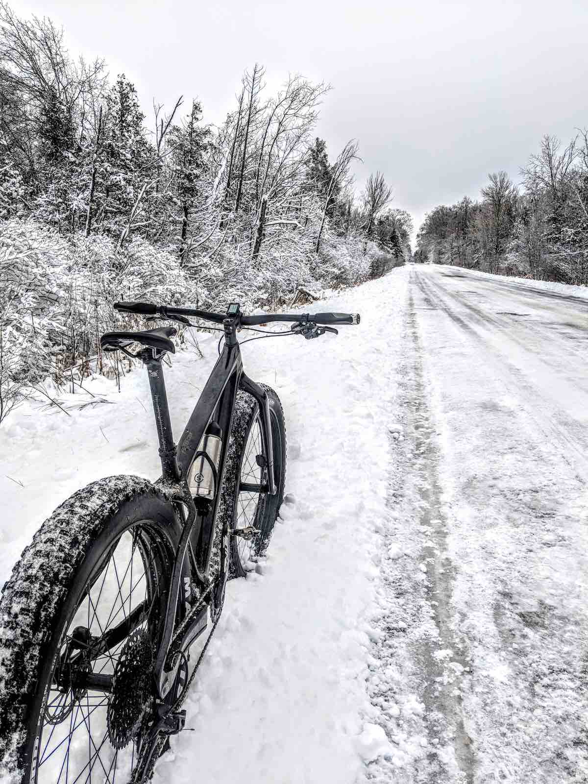 bikerumor pic of the day winter in cadillac, michigan. black mountain bike on the side of a snowy road with trees covered in snow on the sides.