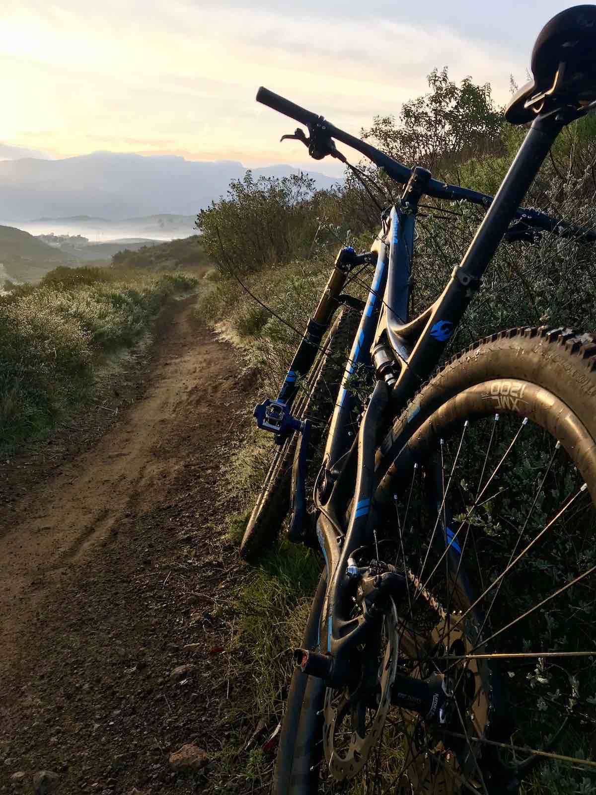 bikerumor pic of the day open trail on the Dos Vientos trail in california, right after sunrise. Mountain bike leaning against berm on dirt trail as morning light fills the sky.