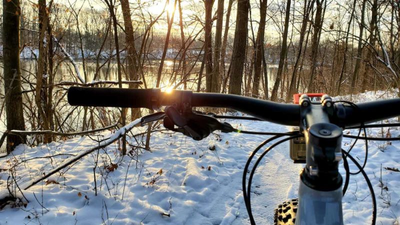 bikerumor pic of the day sunrise in Anderson park Trail in Lansing, MI. view from cockpit of a mountain bike looking over snowy trail as sun rises through trees over the lake.