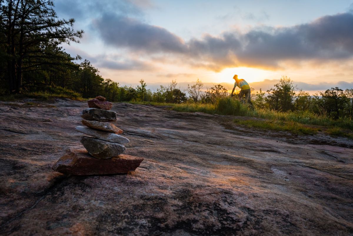 bikerumor pic of the day dear rock, dupont state park north carolina. mountain biker is in the distance riding on slick rock with the sun setting behind them and a cairn in the foreground.