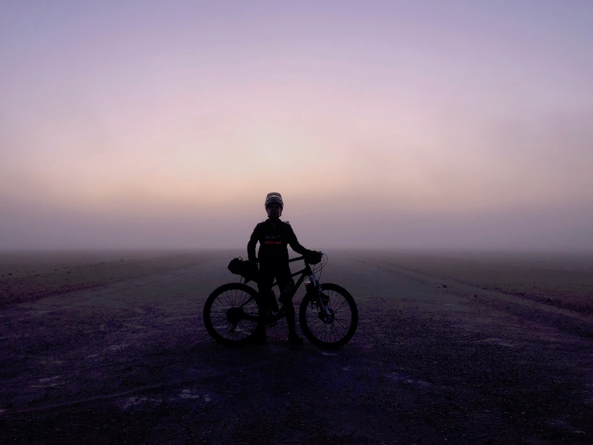 bikerumor pic of the day cycling through the namib desert in namibia. photo is the figure of a cyclist standing in the desert. ground and cyclist are dark with a purple haze in the sky as the sun rises.