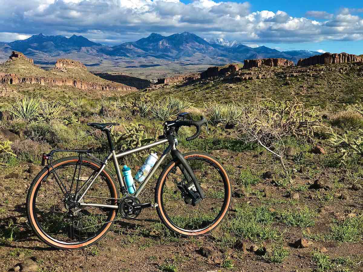 bikerumor pic of the day moots bicycle posed among the scrub on monolith garden trail in kingman arizona, rocky mountain ridge in the distance with blue skies and heavy white clouds above them.