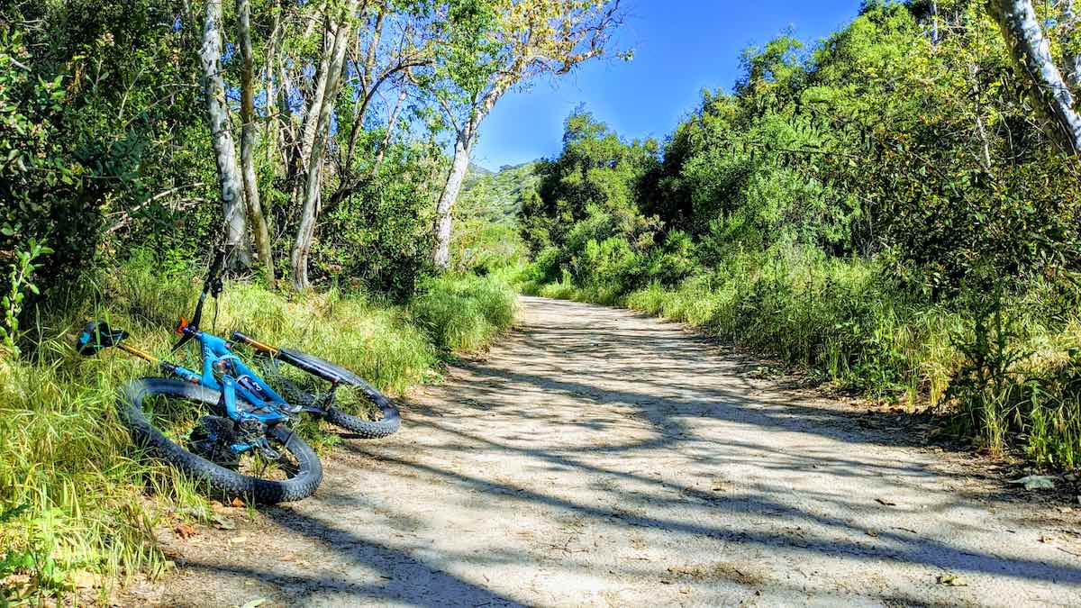 bikerumor pic of the day Laguna Canyon Wilderness Park, Laguna Beach, CA, blue mountain bike leaning on the side of a hard packed path with green trees and brush on either side.