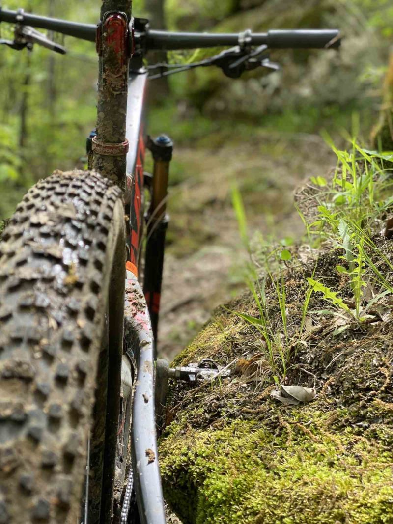 bikerumor pic of the day pivot mountain bike with mud on the tires leaning up against a mossy berm with new grass and leaves in the background.