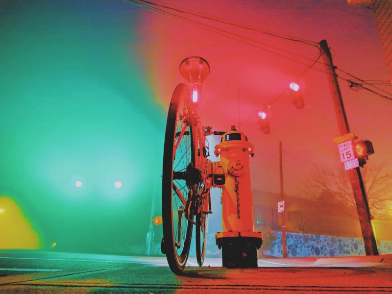 bikerumor pic of the day foggy morning commute in covington kentucky, a bicycle leaning against a fire hydrant by the street with fog making the street lights glow green on one side of the photo and red on the other side.