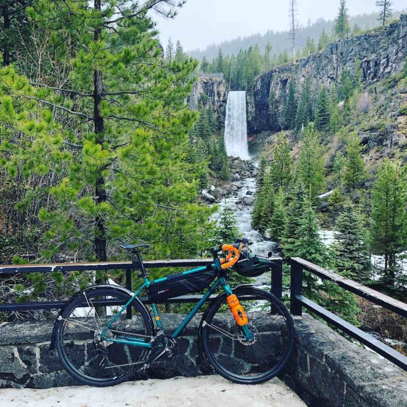 bikerumor pic of the day touring bike in leaning against a railing overlooking a waterfall and pine trees.