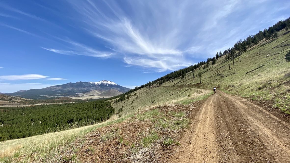 bikerumor pic of the day saddle mountain in norther arizona bicycle on a dirt road on the grassy side of a mountain with a snowy peak in the distance.