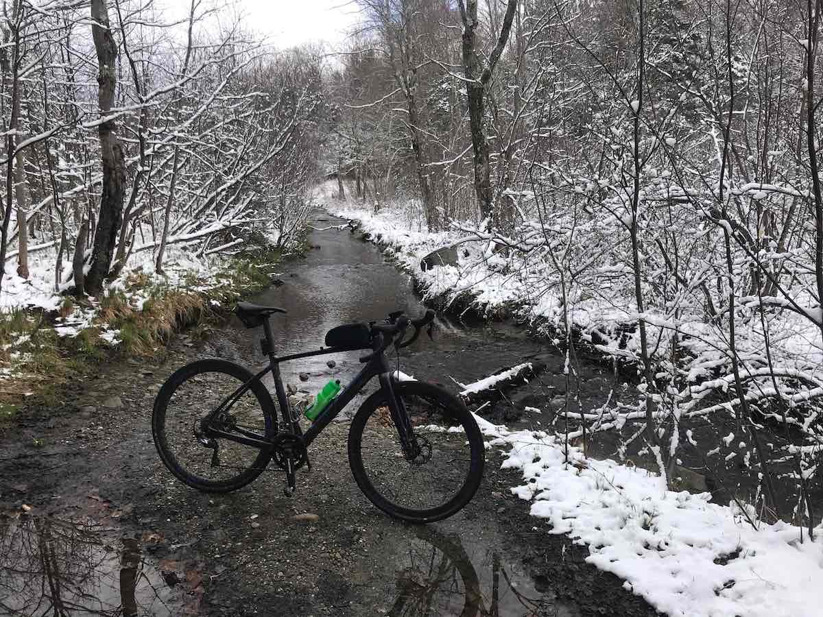 bikerumor pic of the day black bicycle in the middle of a roadbed with creek water running into it snow on the banks and covering leafless trees in jeffersonville vermont.