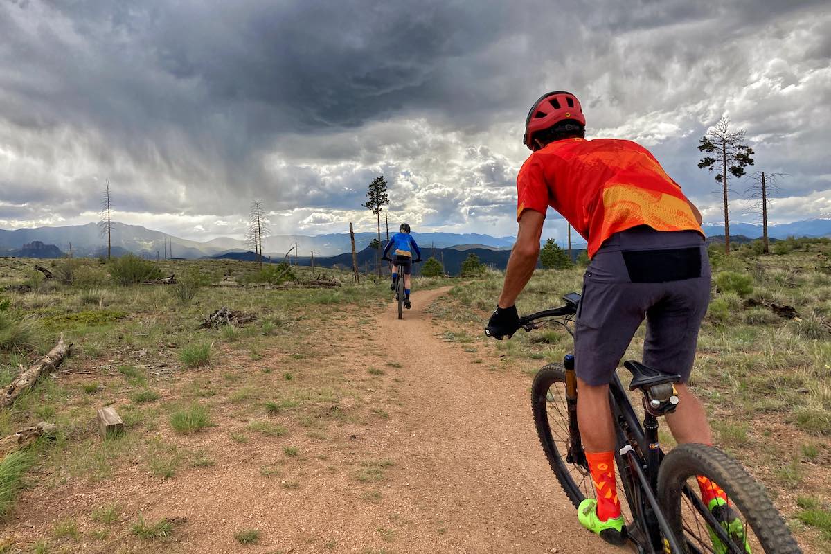 bikerumor pic of the day two mountain bikers on a flat dirt path with greens scrub bushes and dark clouds in the sky.