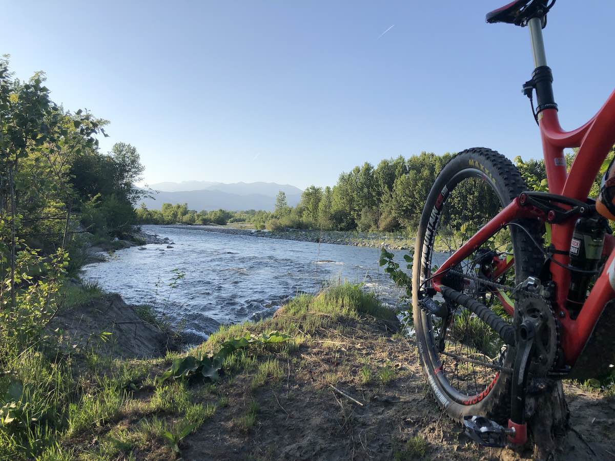 bikerumor pic of the day rear wheel of a bicycle on the edge of a river with green trees on the banks, low mountains in the distance.