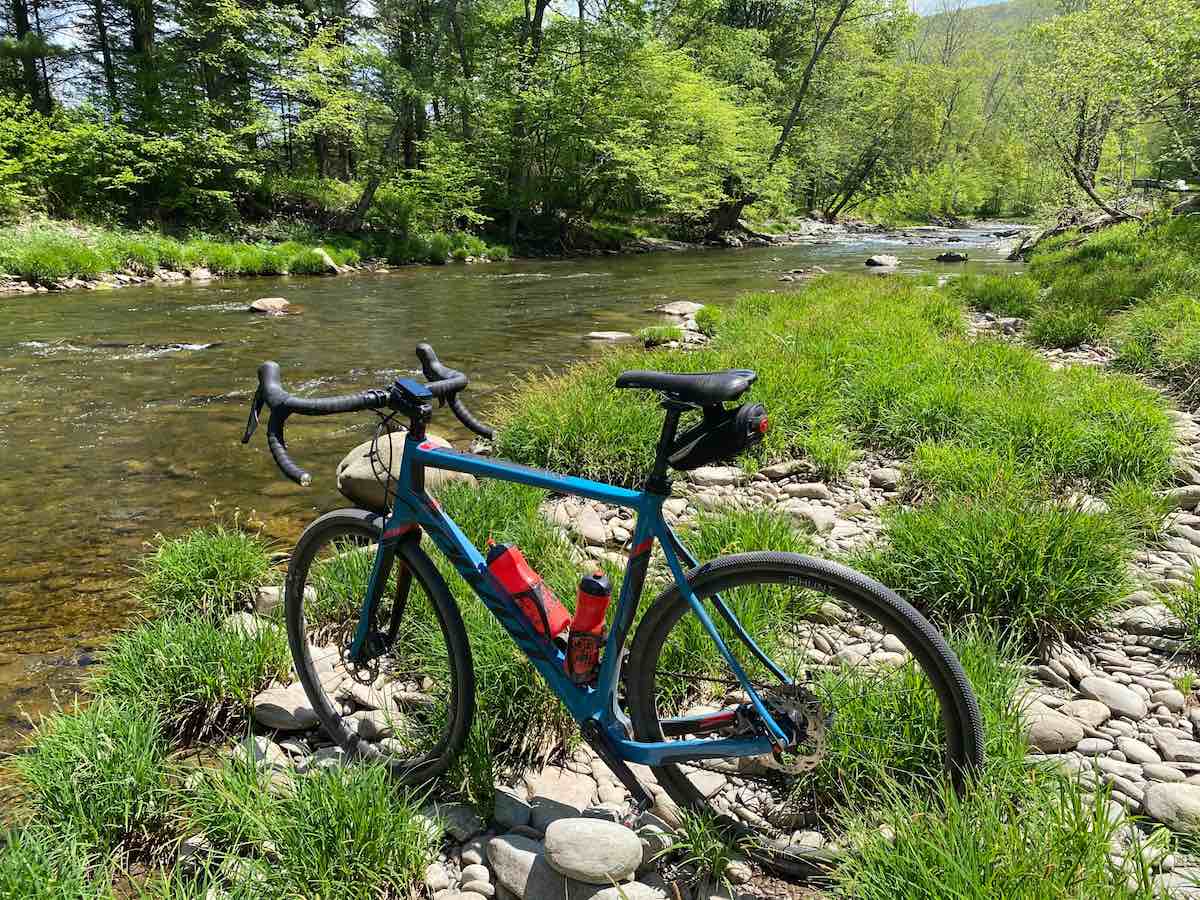 bikerumor pic of the day pivot bicycle in the grass and rocky side of a creek with lots of green trees in the distance.