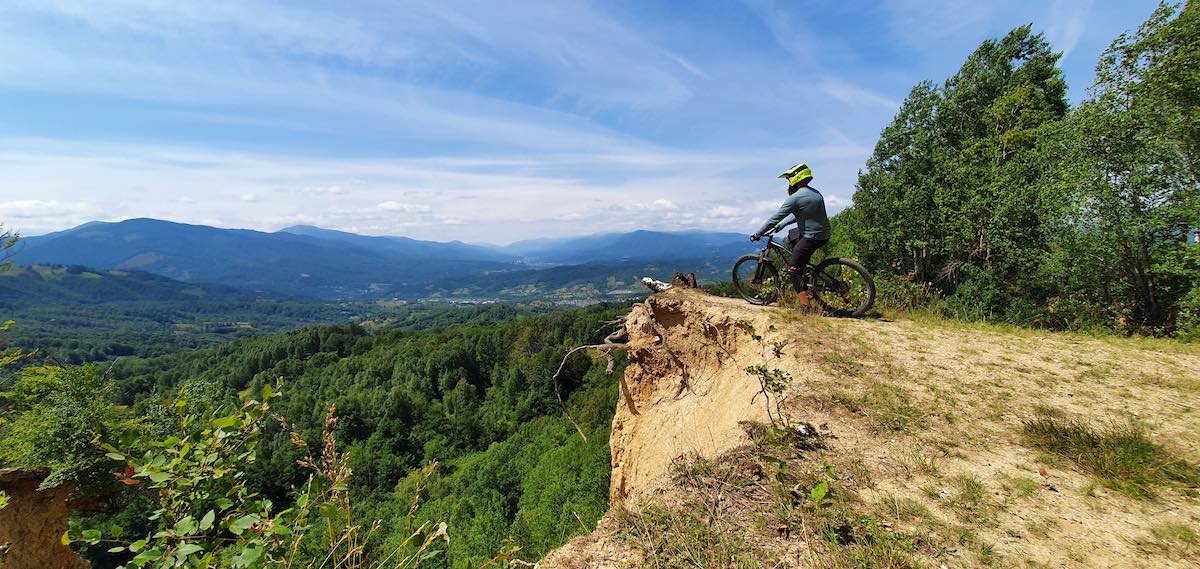 bikerumor pic of the day parang mountains romania cyclist sits on a mountain bike on the edge of a small cliff overlooking green forest and a mountains beyond.