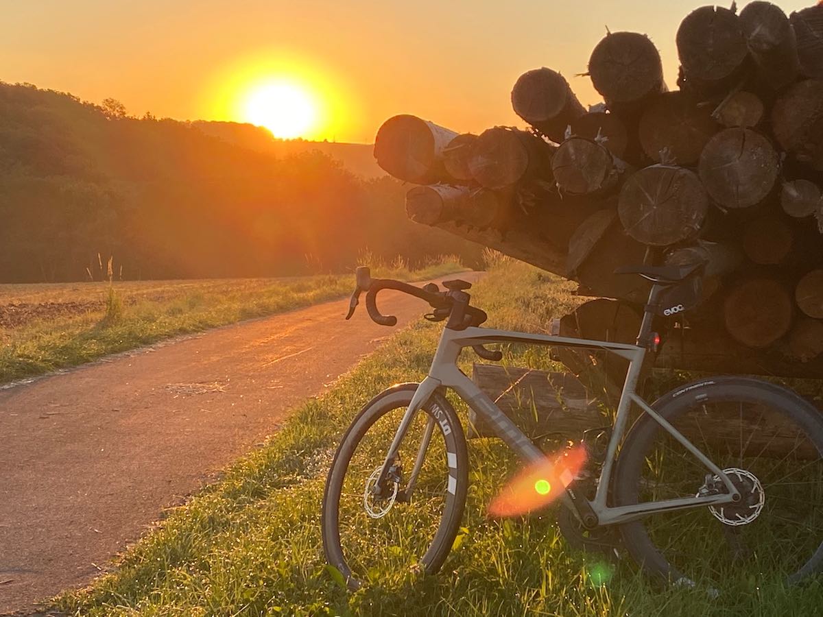 bikerumor pic of the day bmc bike leaning against a pile of logs alongside a path with the orange glow of the sunset in the distance.