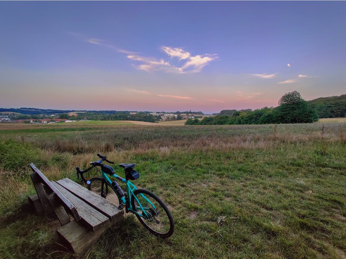 bikerumor pic of the day cycling near düsseldorf germany, teal bicycle leans against a wooden bench overlooking a wide field with a low orange sky at dusk