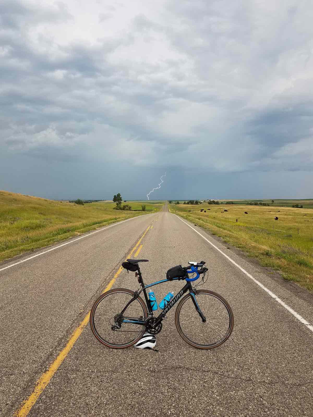 bikerumor pic of the day a specialized road bike is posed in the middle of a long straight road with flatlands on either side and a dark storm with one lone lightning strike in the distance.