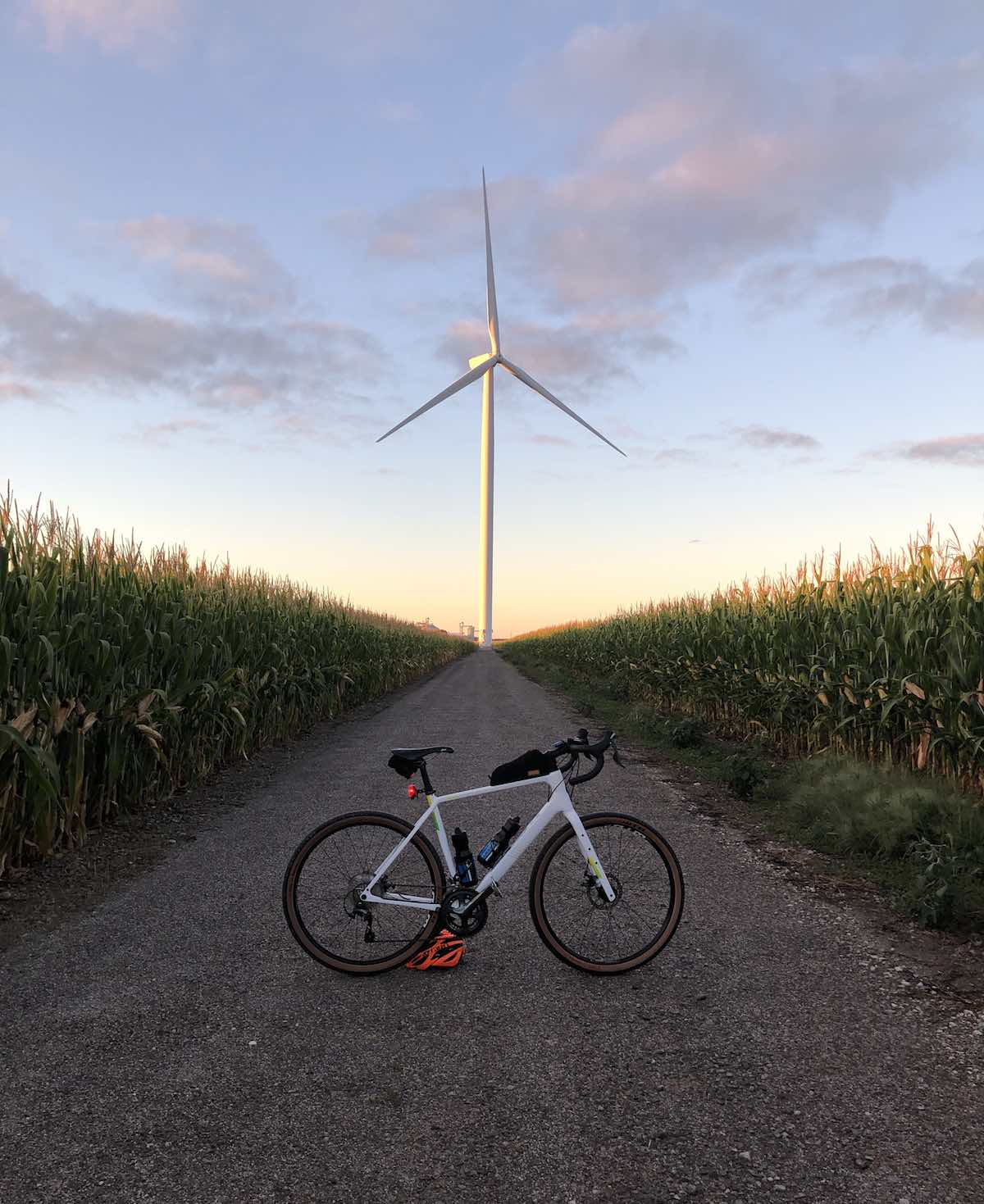 bikerumor pic of the day bicycle in the middle of a gravel road with corn growing on both sides leading to a modern windmill at dusk