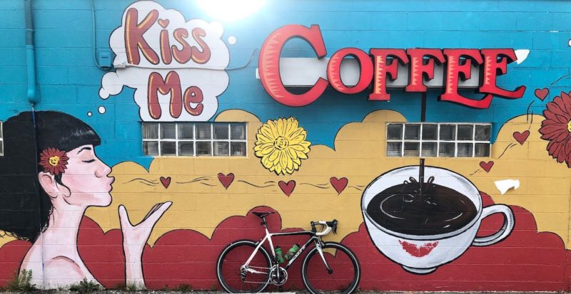 bikerumor pic of the day bicycle posed in front of a mural for a coffee shop called Kiss Me Coffee showing a woman with white skin and black hair blowing kisses to the coffee cup.