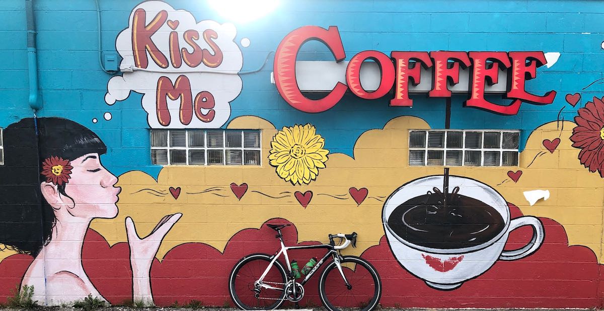 bikerumor pic of the day bicycle posed in front of a mural for a coffee shop called Kiss Me Coffee showing a woman with white skin and black hair blowing kisses to the coffee cup.