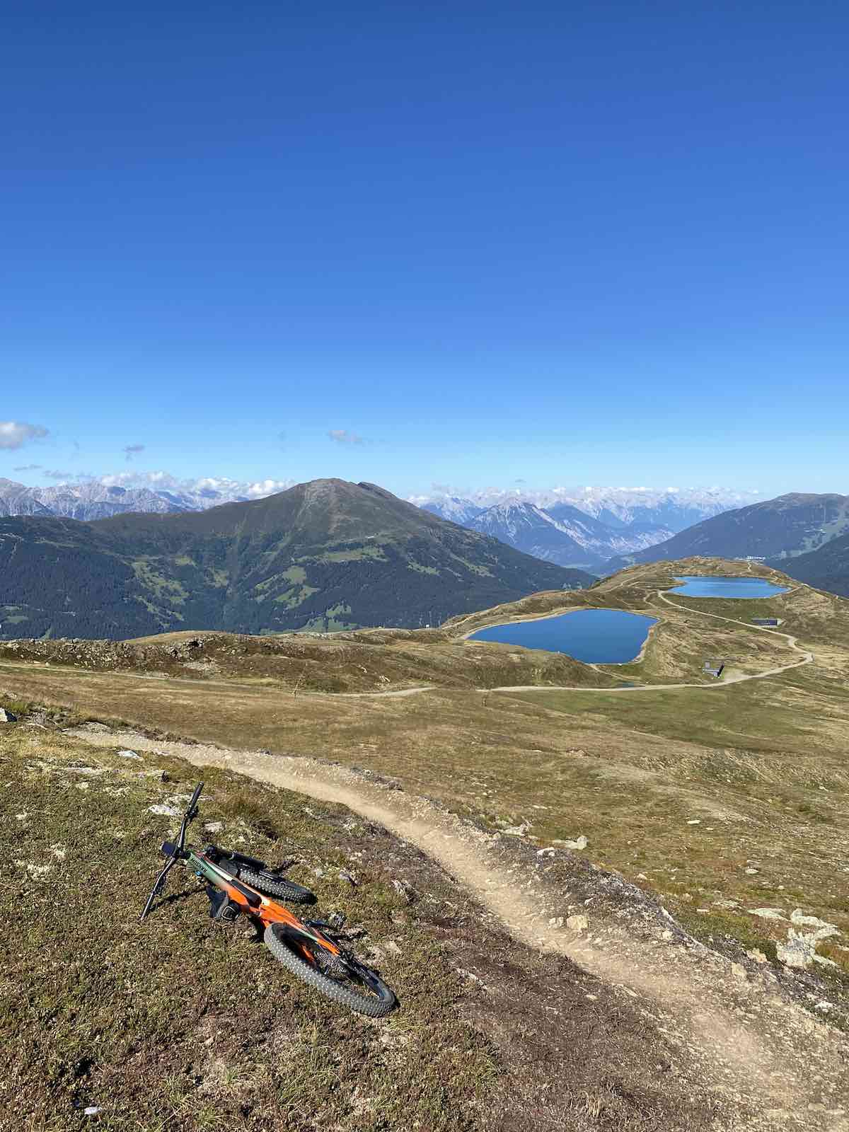 bikerumor pic of the day orange mountain bike is laying beside a dirt trail on a rocky plain leading up to a lake and snow capped mountains beyond with clear blue skies in the distance.