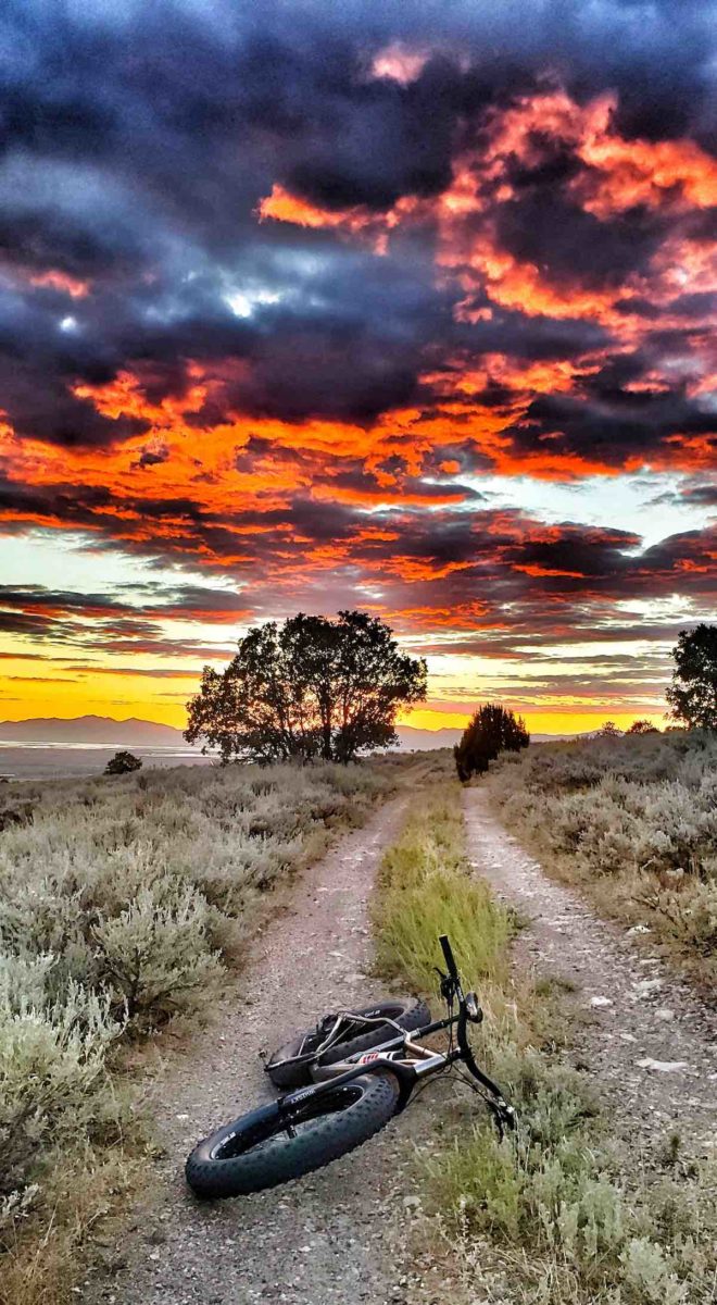 bikerumor pic of the day Ogden Utah fat bike laying on a dirt car path with scrub brush on either side a few sparse trees in the distance and a fiery sky at dusk.