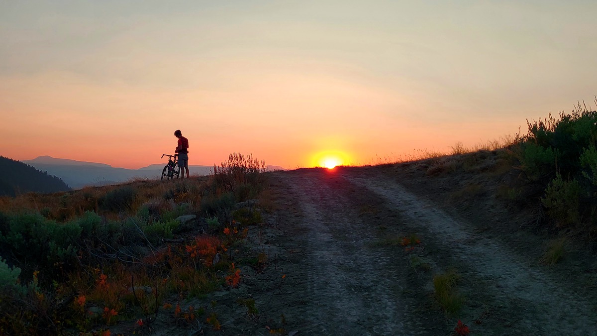 bikerumor pic of the day cyclist stands in relief to the bright orange sun setting in the sky above a dirt road with short plants and scrub brush on either side.
