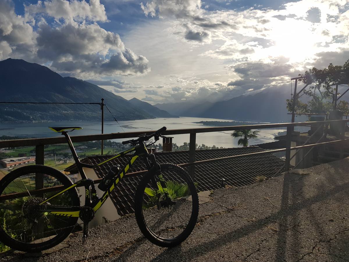bikerumor pic of the day Ticino switzerland scott bicycle leaning against a railing looking out over a town on the edge of a lake with mountains and fluffy clouds in the distance.