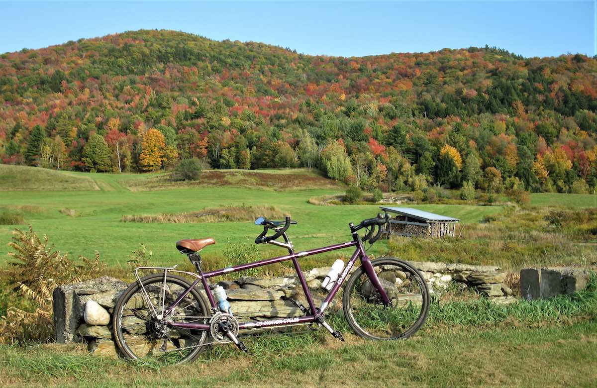 bikerumor pic of the day co-motion tandem bicycle in a grassy field by a low stone wall with mountains in the background that have trees in green gold and red as the fall leaves change.