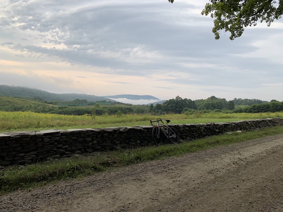 bikerumor pic of the day dummerston vermont bicycle leaning against a low stone wall next to a gravel road and with a green field on the other side of the wall with low tree line and mountains with fog on them in the distance.