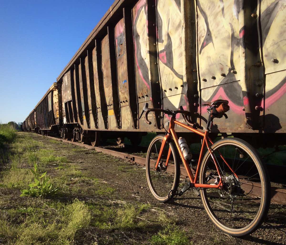 bikerumor pic of the day bicycle next to a stopped train with graffiti on it clear blue skies and green grass.