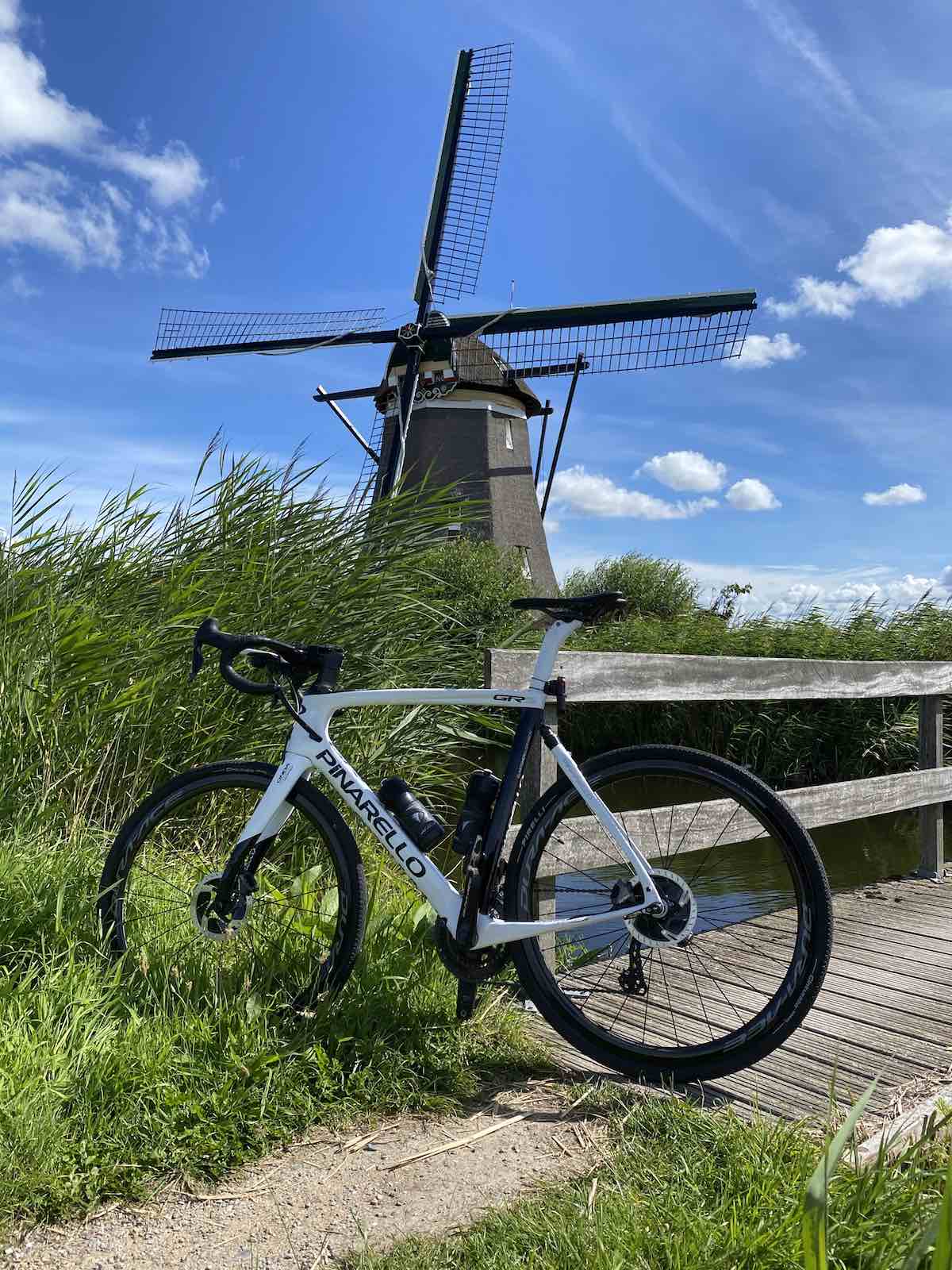 bikerumor pic of the day road bicycle leaning against a wooden bridge with high green grass on either side and a windmill in the distance with bright clear blue sky.