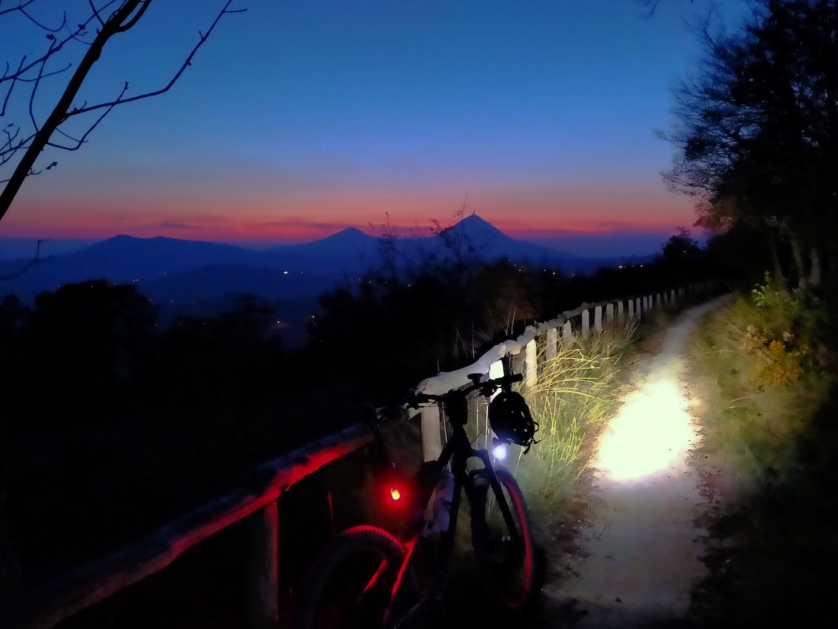 bikerumor pic of the day bike riding at sunset with bike light shining on the path ahead with dark mountain profile and orange red sunset