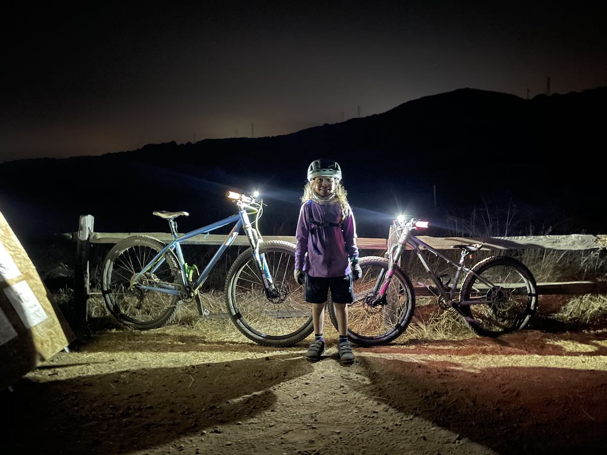 bikerumor pic of the day a small child stands between to mountain bikes that have lights on the handlebars it is dark outside withe the shadow of a mountain in the background and the lights from the bicycles lighting the child from behind.