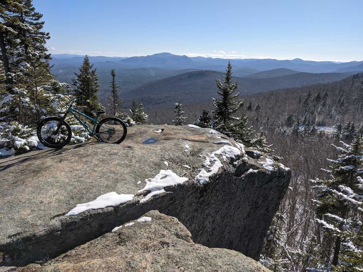 bikerumor pic of the day mountain bike on a large boulder with bits of snow on it overlooking pine trees and mountains in the distance of adirondack park in new york