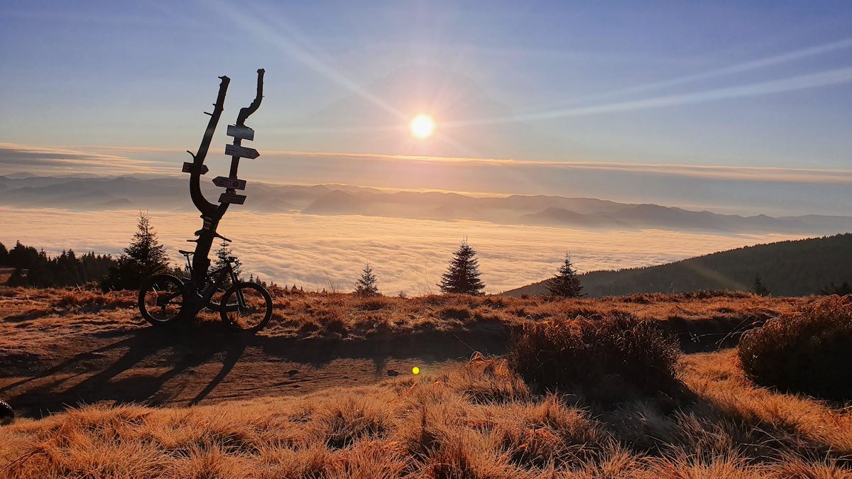 bikerumor pic of the day Martinske Hole, Slovakia a mountain bike leans against a dead tree trunk as the sun sets on golden brush along the top of a mountain with the clouds below.