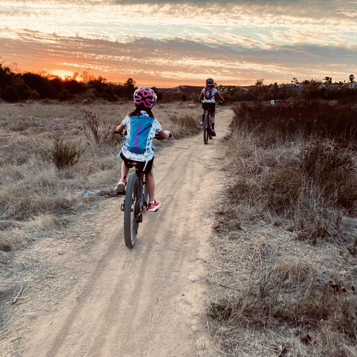 bikerumor pic of the day Los Penasquitos Canyon - San Diego, CA a child and another person on mountain bikes ride on a dirt path with sage colored brush on either side towards a grey and orange sunset.