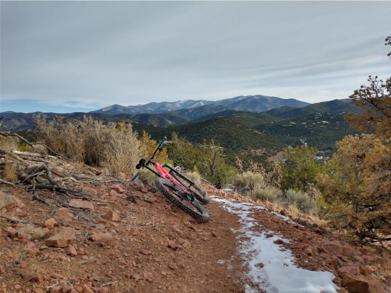 bikerumor pic of the day dale ball trails santa fe new mexico a bicycle lays on the side of a dirt and rocky trail with a bit of snow sage brush is on the side with dark green rolling mountains in the distance.