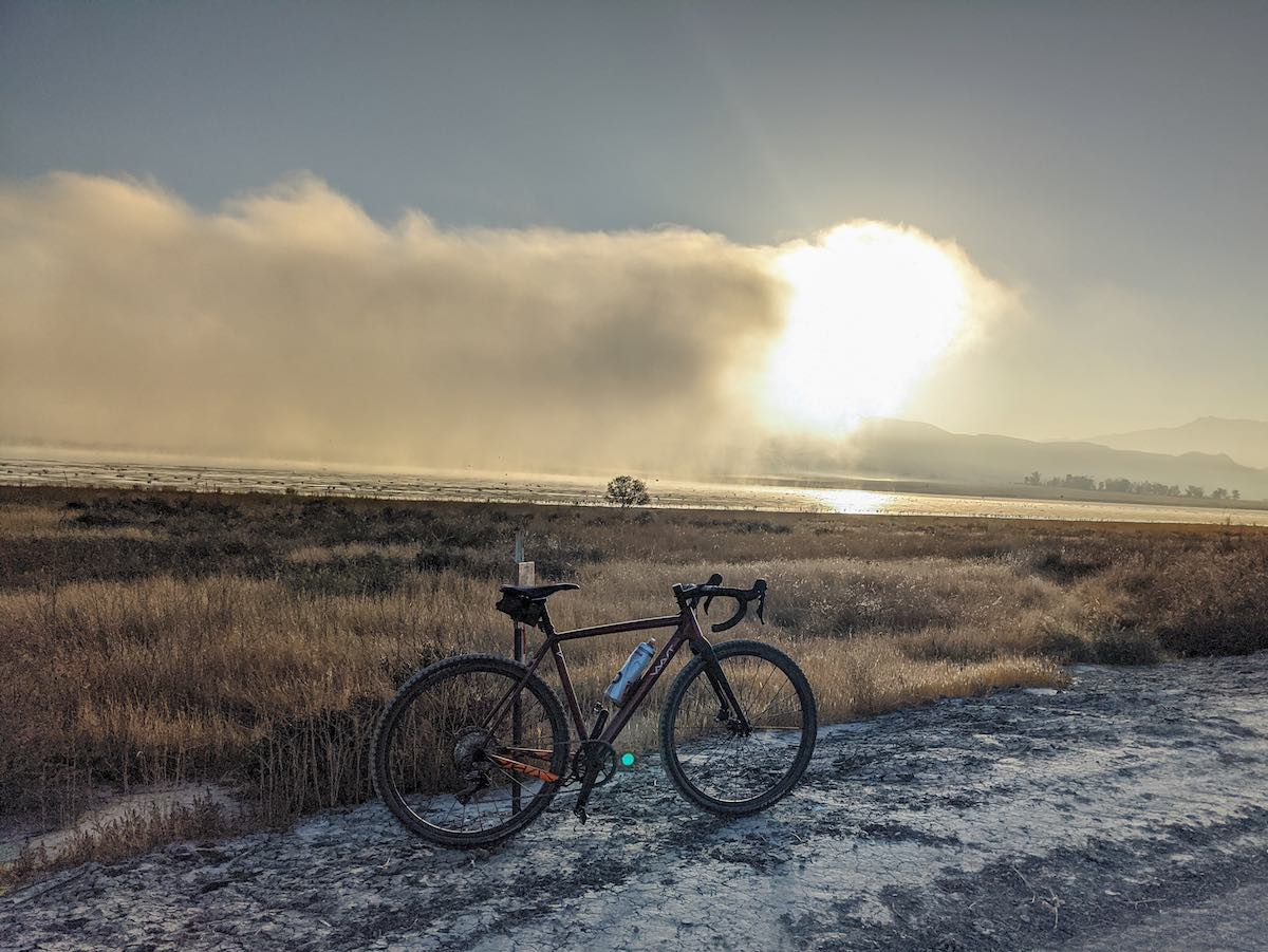 bikerumor pic of the day a vaast a/1 bicycle is posed on a dirt road with mystic lake in the distance. the sun is low and it looks like fog is just burning off the lake.
