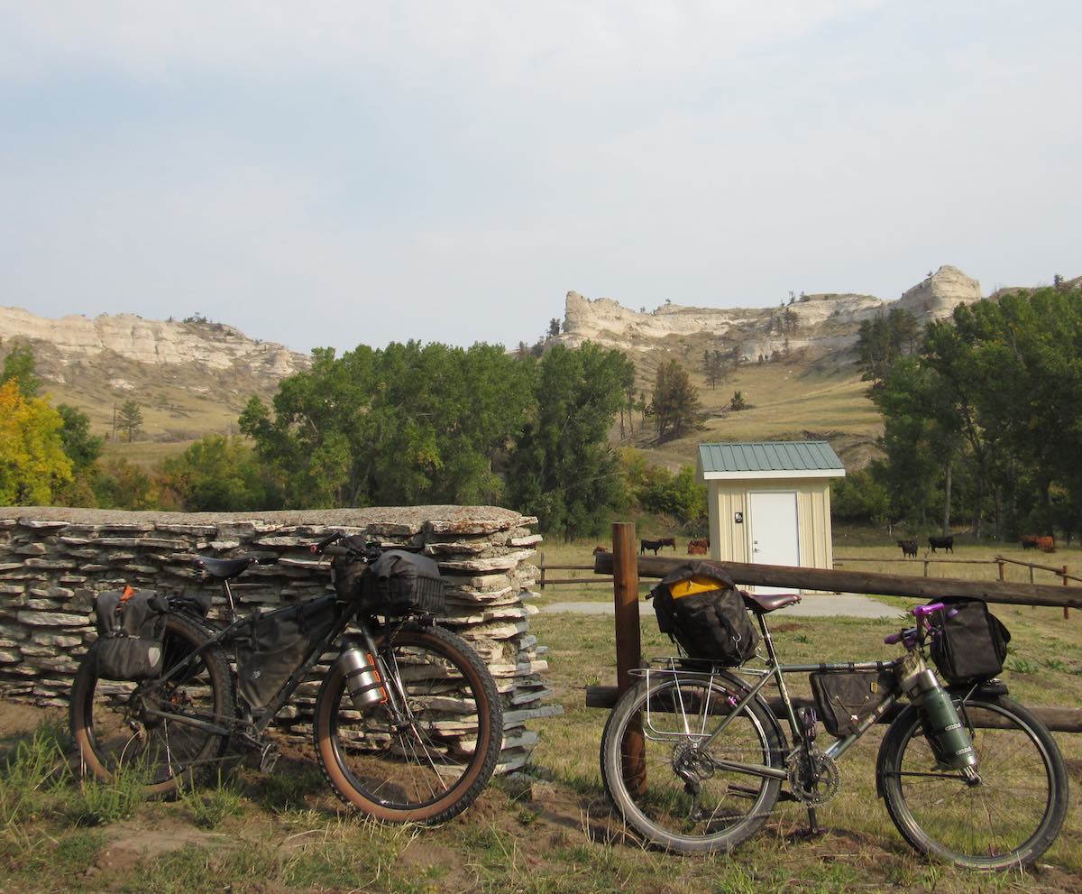 bikerumor pic of the day bikepacking in harrison nebraska. trek 820 mountain track and rawland leaning against a stone and wood fence with cows in the distance and a mountain feature sticking out above the green trees.