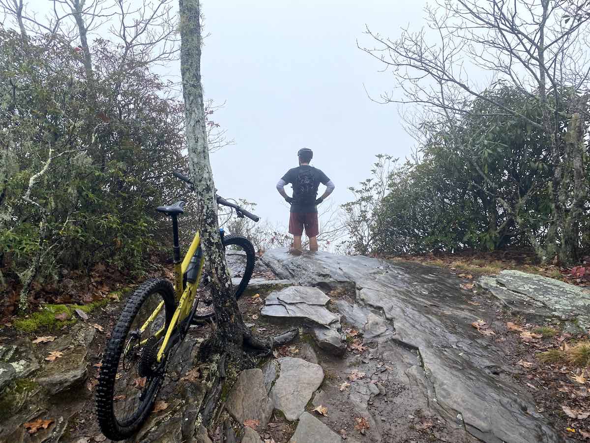 bikerumor pic of the day a cyclist stands at the edge of a slick rock looking into the cloud that is covering the mountain with a yellow mountain bike leaning against a skinny tree in the foreground at pisgah national forest in brevard north carolina.