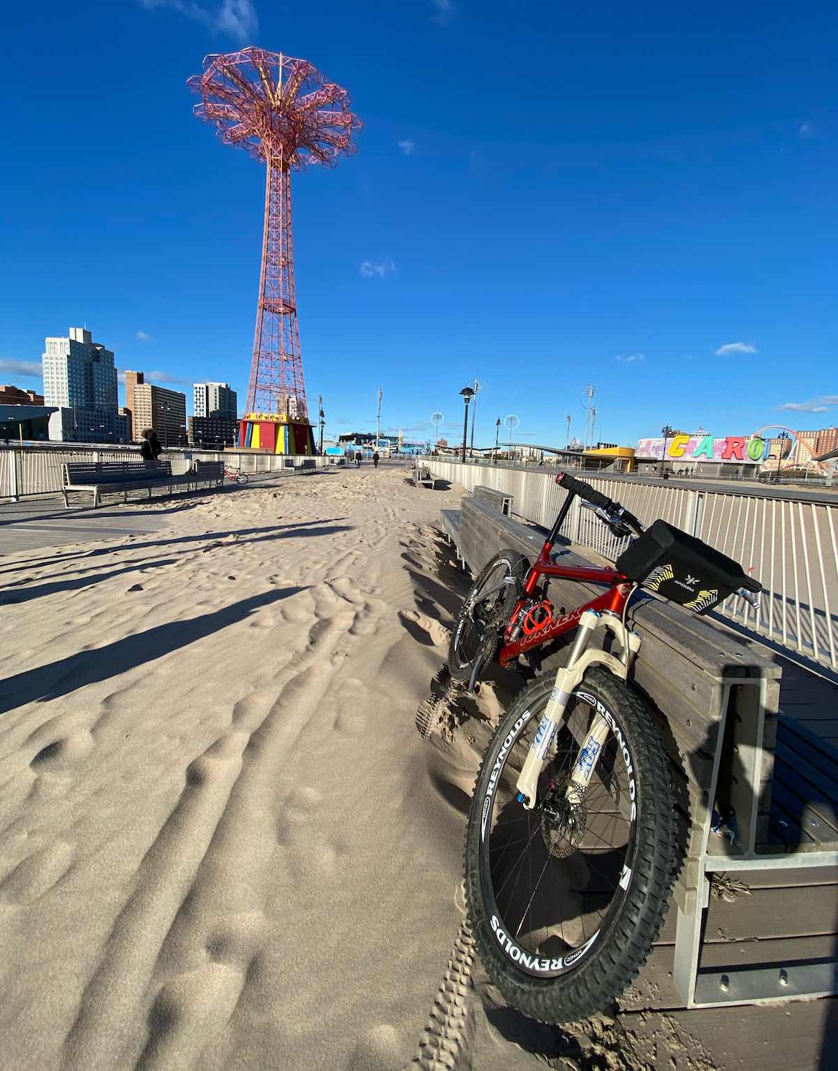 bikerumor pic of the day a mountain bike leans against a bench on the boardwalk of coney island there is sand covering the boardwalk and carnival rides in the distance, it is a clear bright winter day and the shadows are long.