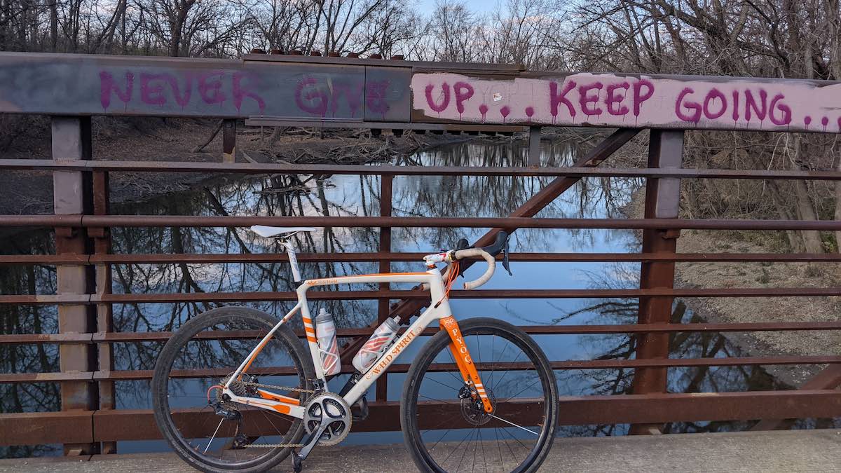 a white and orange road bike leans against the metal railing of a bridge over a body of water with graffiti on the metal beam above that says never give up, keep going
