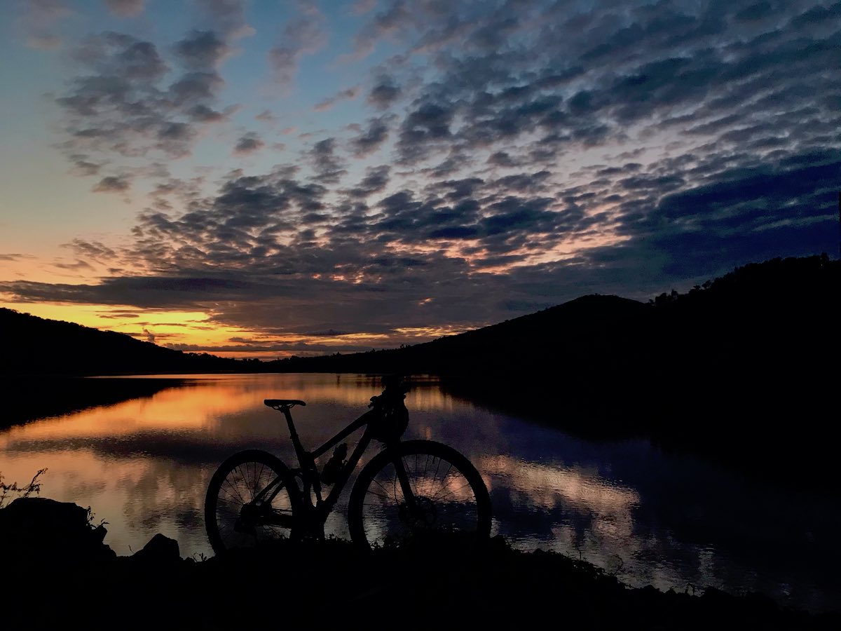 bikerumor pic of the day a bicycle is in profile parked before a body of water with the reflection of the morning sun peeking through dark clouds