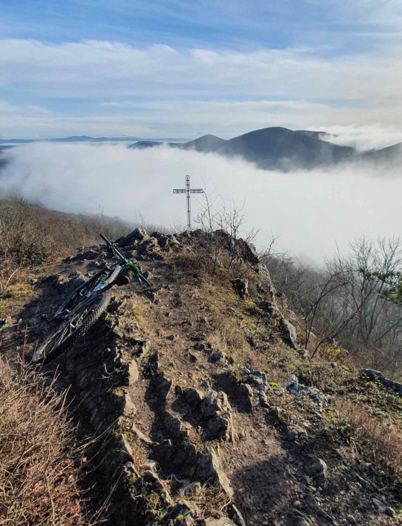 bikerumor pic of the day mountain biking in slovakia a bicycle is leaning against a rocky outcrop so high in the mountains it is above the clouds, a cross is seen in the distance.