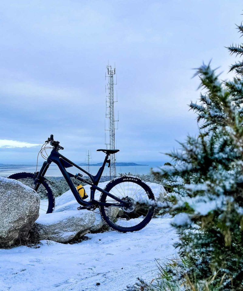 bikerumor pic of the day Ticknock Mountain in dublin ireland, a canyon spectral mountain bike leans against a large rock next to a path covered in snow a pine tree to the right of the pic and cell tower in the distance.